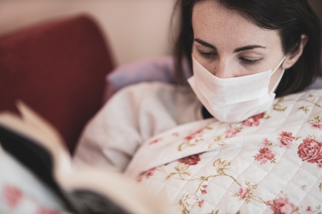 Femme malade avec un masque chirurgical entrain de lire dans son canapé sous un plaid